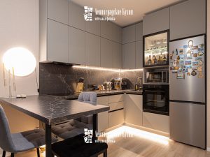 Современная угловая кухня в с барной стойкой с матовыми фасадами серого цвета с системой открывания push to open с оригинальной черной столешницей и фартуком с текстурой камня с золотыми прожилками, подсветкой зоны приготовления и обеденной зоны